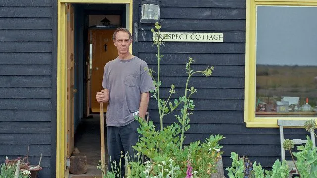 Derek Jarman, en la fotografía, aparece como un hombre alto y delgado de mediana edad con pelo cano y alborotado. Lleva una camiseta vieja de color gris. Aparece frente a la fachada de la casa, de tablas de madera pintadas de negro y adornos en amarillo. El cartel que hay junto a la puerta muestra el nombre de la cabaña, Prospect Cottage.