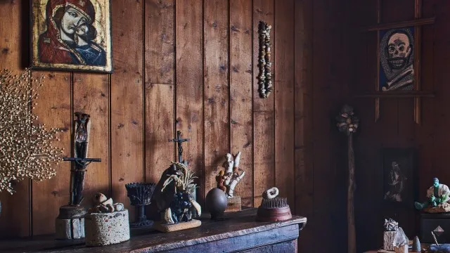 Una pared del interior de la cabaña, hecha de tablas verticales de madera oscura. Hay un cuadro colgado que parece ser un icono de la virgen María acompañando al niño Jesús, de estilo eslavo.  Sobre un mueble, pequeñas figuras de madera, cuerda, piedras y ramitas sin formas definidas. A la derecha se ve una figura del increíble Hulk y un mineral sobre un pequeño estandarte, como de una colección.